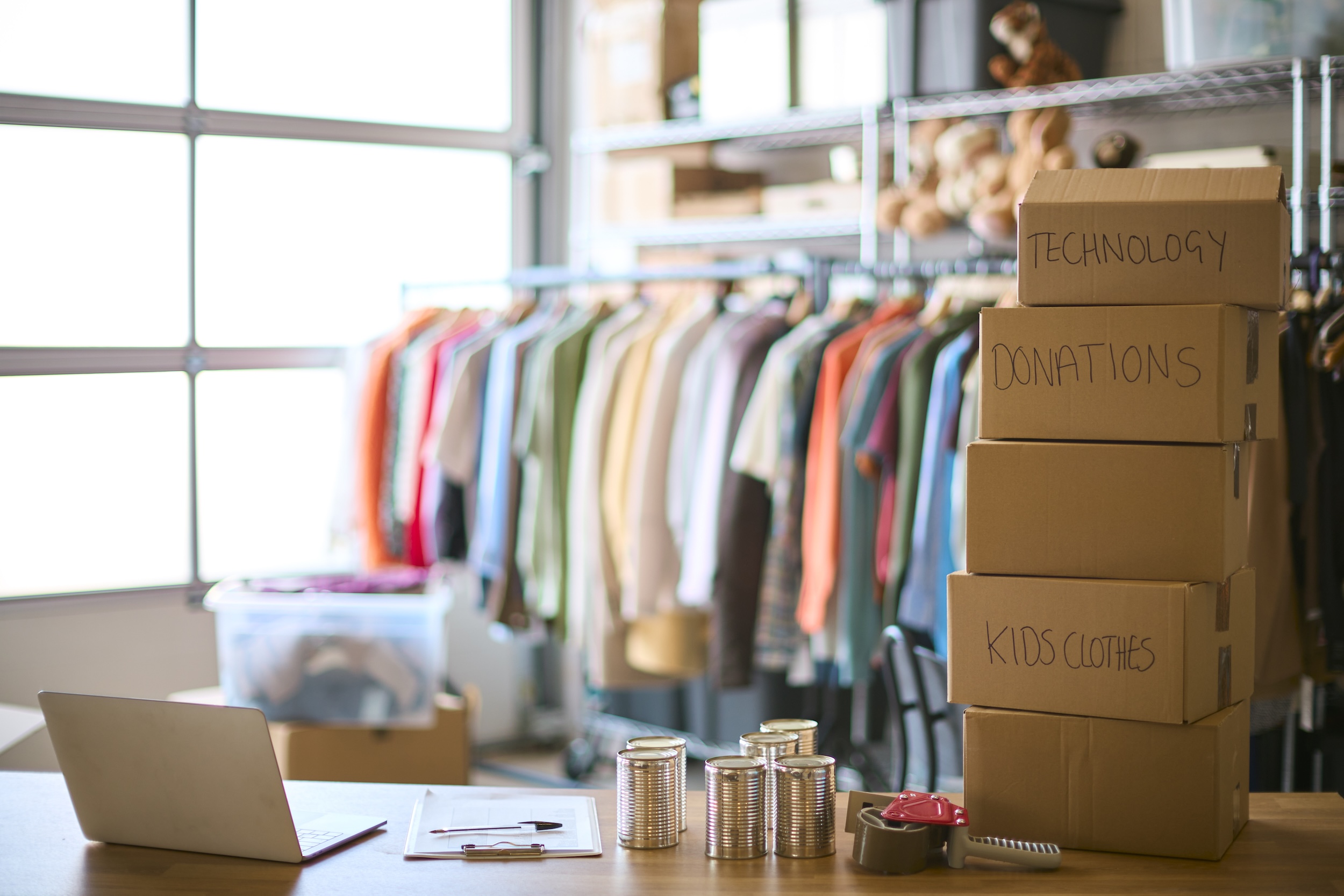 Cajas de cartón apiladas con etiquetas escritas a mano junto a perchero con ropa colorida y portátil sobre mesa en espacio luminoso
